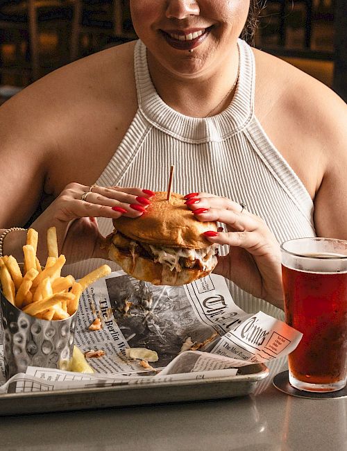Person wearing a white halter top holding a burger with French fries on a tray and a red drink on the side, at a table.