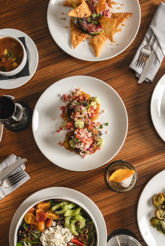 A bird&rsquo;s-eye view of multiple plated dishes on a wooden table: tacos, salads, salmon, stew, and a variety of colorful sides, plus drinks&mdash;delicious feast.