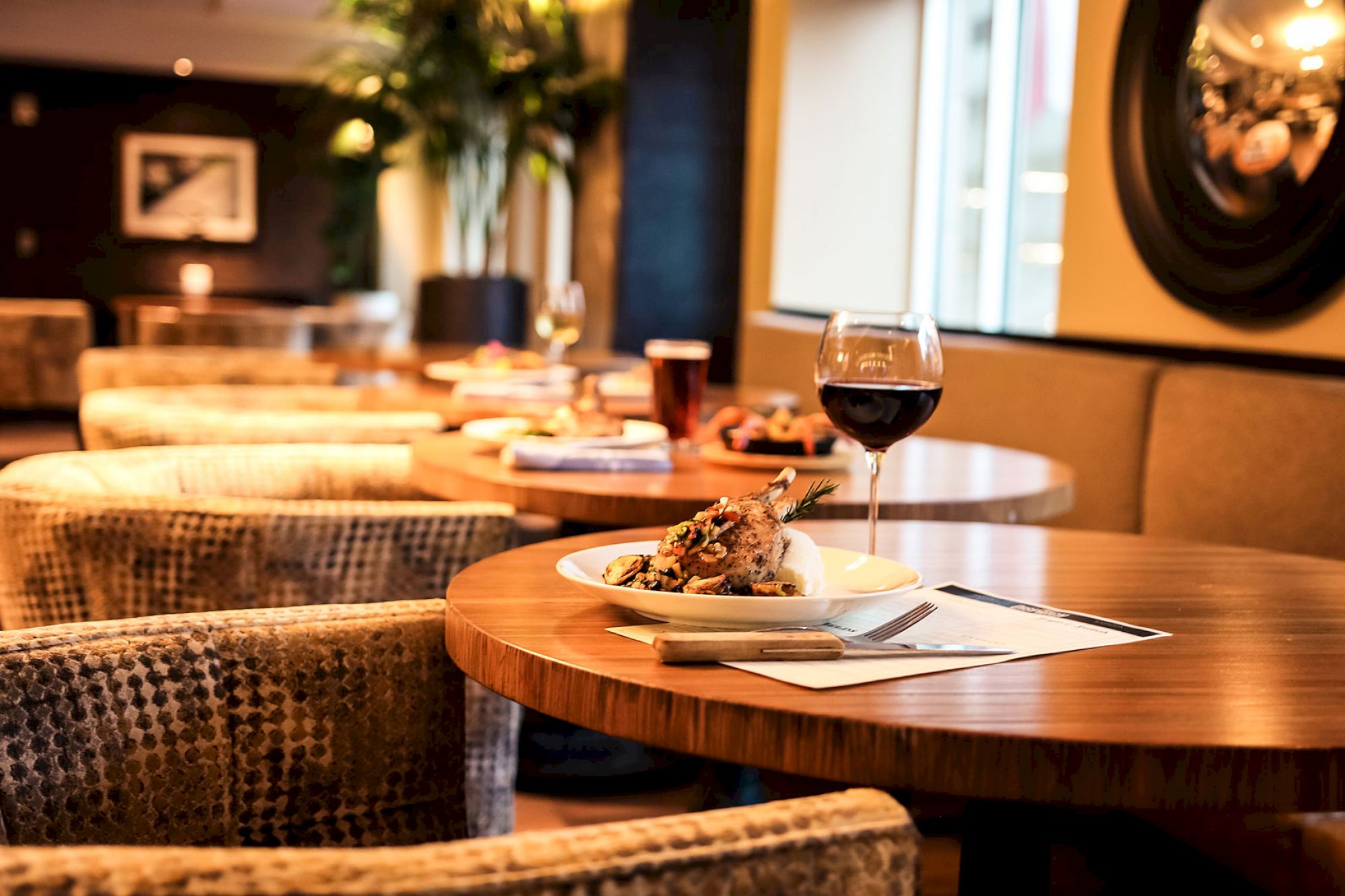 A restaurant table with a glass of red wine and a plate of food, featuring a modern and cozy ambiance with cushioned chairs and tasteful decor.