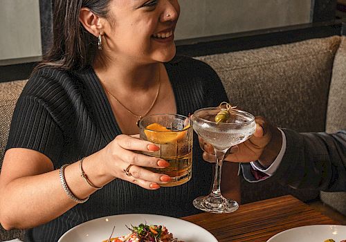 Two people raising drinks and toasting over a table of colorful plated dishes, smiles and clinking glasses, vibrant food scene, cheers.