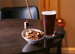 A glass of beer and a bowl of snacks on a table, with pool cues and a pool ball in the background.