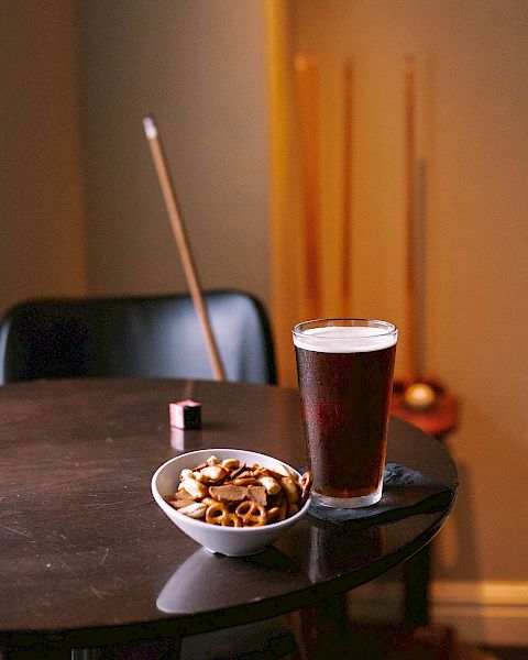 A glass of beer and a bowl of snacks on a table, with pool cues and a pool ball in the background.