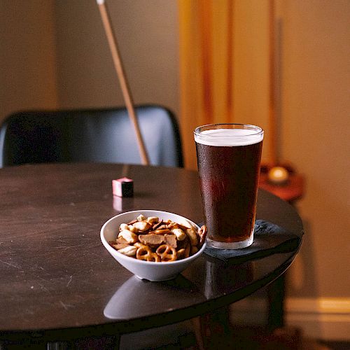 A glass of beer and a bowl of snacks on a table, with pool cues and a pool ball in the background.