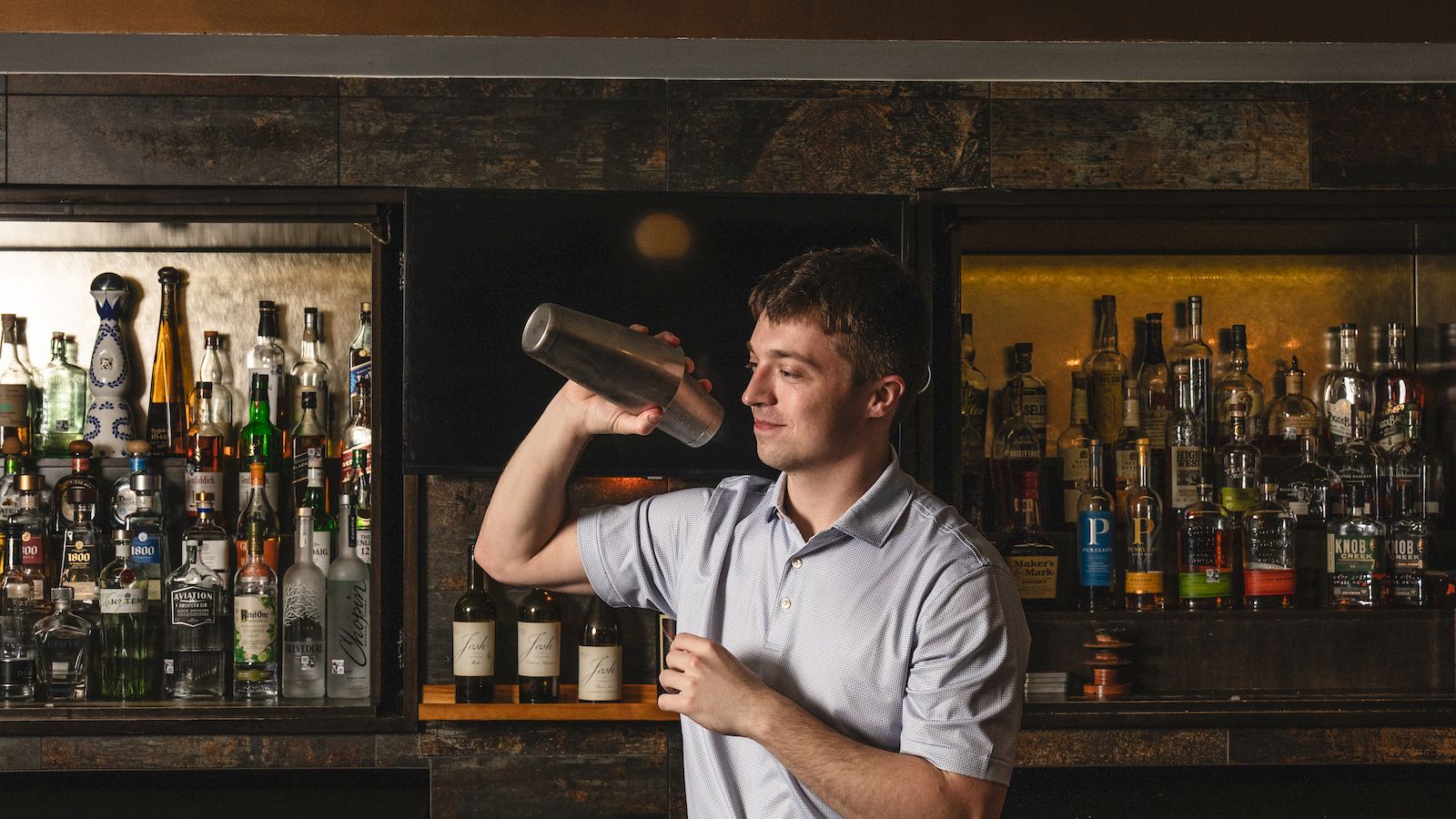 A bartender shakes a drink behind a dim, vintage bar as bottles line the shelves; warm light casts a cozy, late-evening glow.