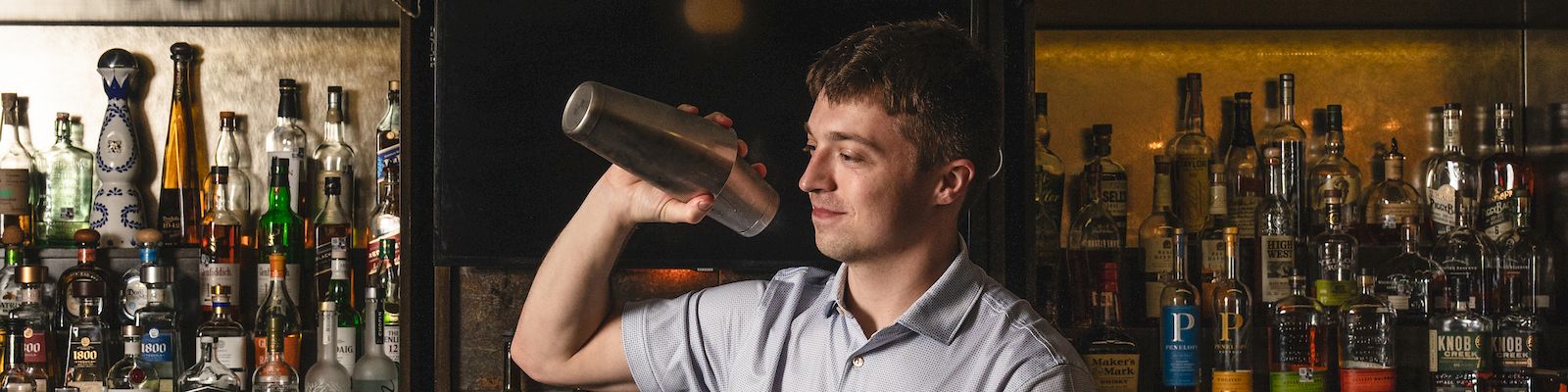 A bartender shakes a drink behind a dim, vintage bar as bottles line the shelves; warm light casts a cozy, late-evening glow.