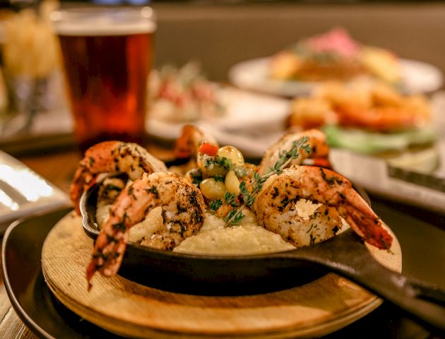 The image shows a skillet with shrimp and some garnish, accompanied by various dishes and a glass of beer in the background.