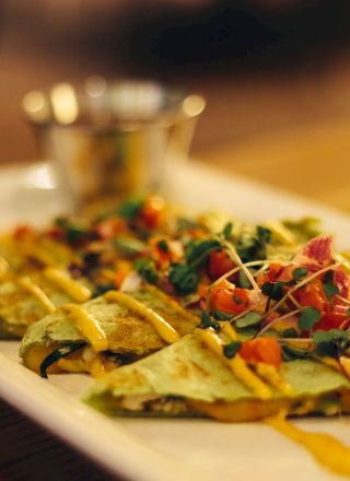 The image shows a plate of quesadillas topped with sauce and garnished with microgreens and diced vegetables, served with a side dip.