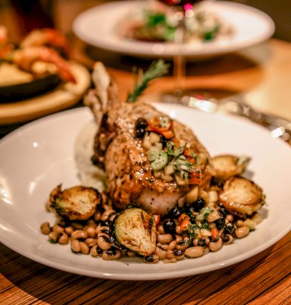 A dish consisting of a cooked meat on a bed of beans and roasted vegetables, with another plate of food in the background, on a wooden table.