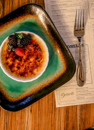 A dessert garnished with berries on a green plate, set next to a fork and menu on a wooden table, plus a drink with an orange slice nearby.