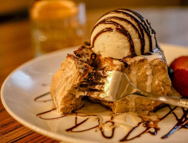 A dessert plate with a fork, featuring a slice of cake topped with ice cream and chocolate drizzle, plus a strawberry garnish in the background.