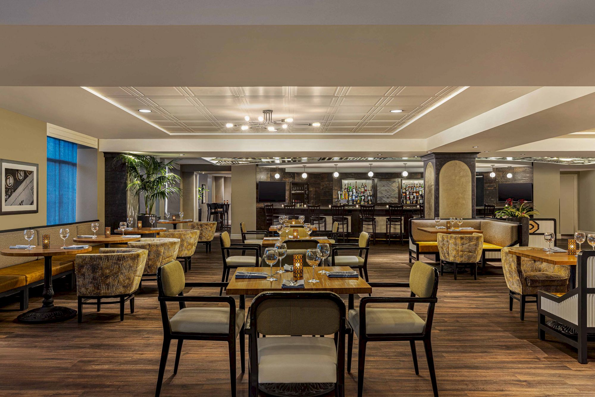 This image shows a modern restaurant with wooden flooring, elegantly set tables, upholstered chairs, and a well-stocked bar in the background.