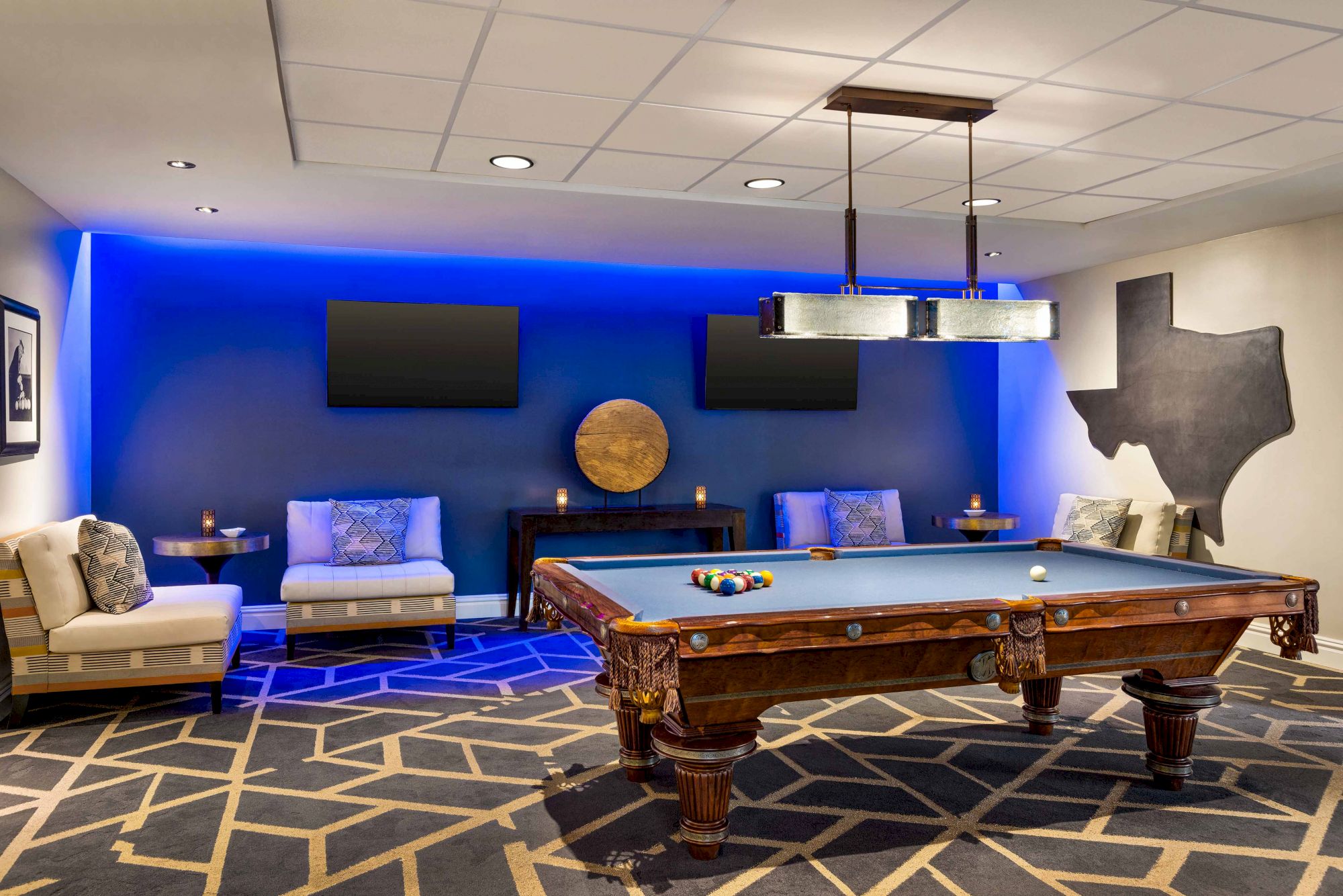 The image shows a stylish game room with a billiard table, two couches, a geometric-patterned carpet, and TVs on a blue-lit wall.