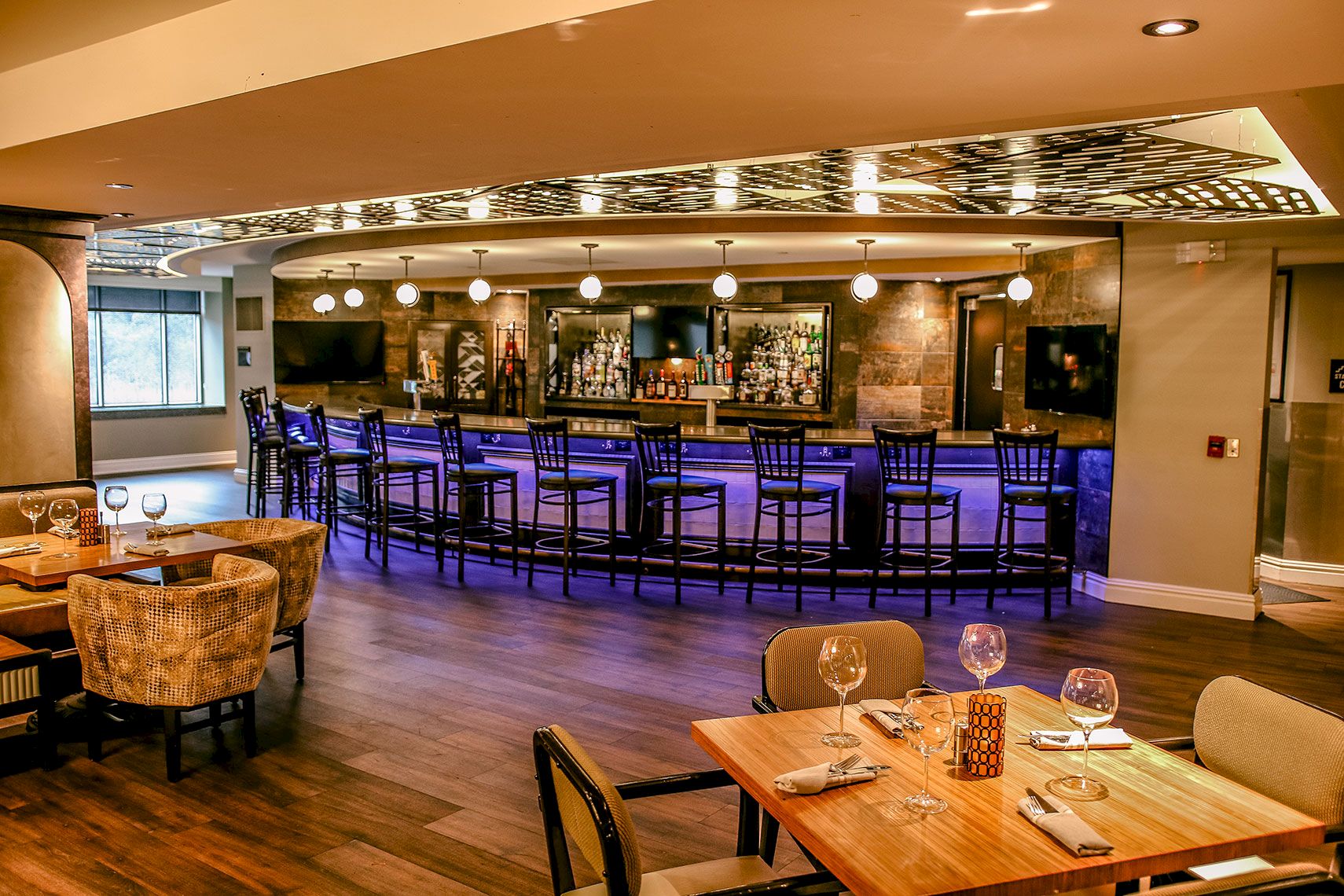 This image features a modern, stylish bar with high chairs and tables set with wine glasses and tableware in the foreground, all under warm lighting.
