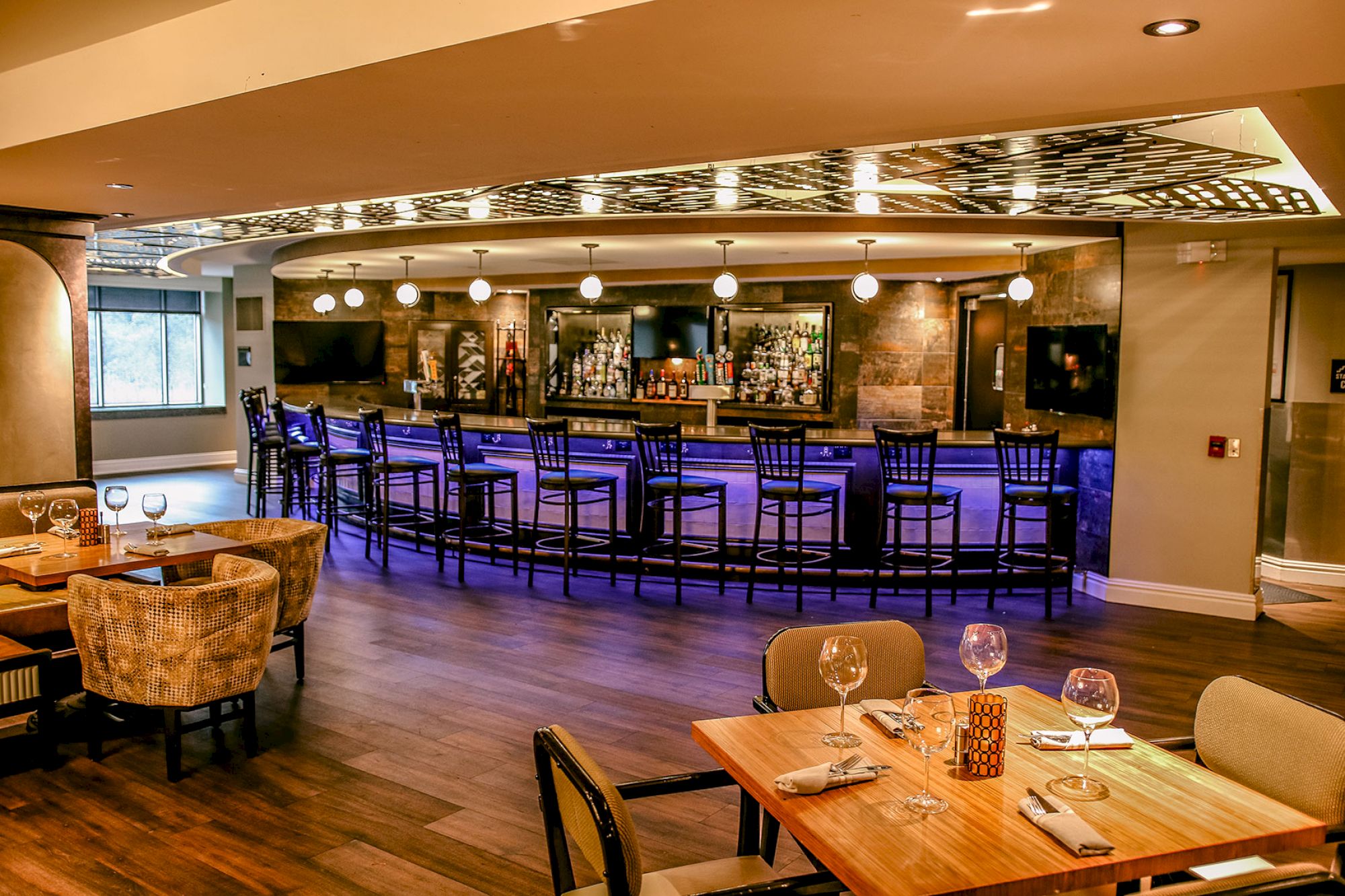 This image features a modern, stylish bar with high chairs and tables set with wine glasses and tableware in the foreground, all under warm lighting.