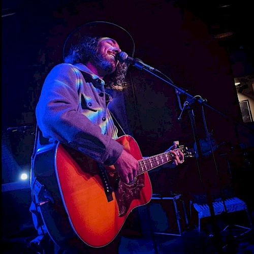 A musician is playing an acoustic guitar and singing into a microphone on a dimly lit stage, wearing a hat and a jacket, under blue lighting.