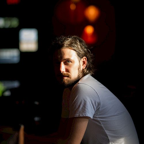 A bearded man in a white T-shirt sits in a dimly lit room with warm lights in the background, gazing at the camera.