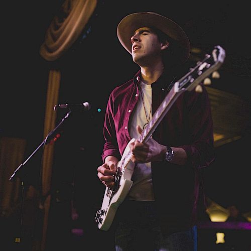 A person wearing a hat is playing an electric guitar, seemingly performing live on stage, with a dark background and some lights behind them.
