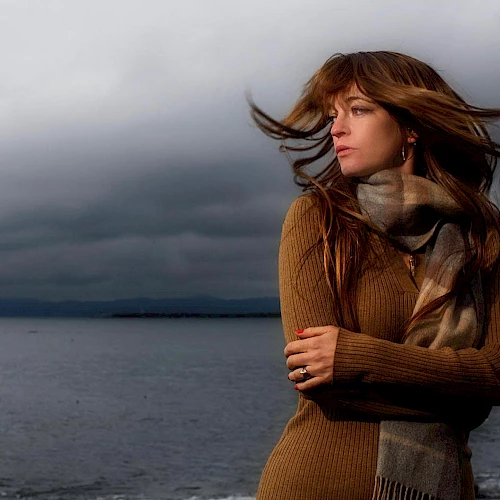 A woman stands by a body of water with a stormy sky in the background, wearing a scarf and sweater, with her hair blowing in the wind.