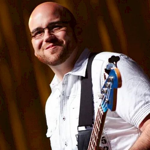 A person is smiling while holding an electric bass guitar. They are dressed in a white shirt and glasses, with a dark background.