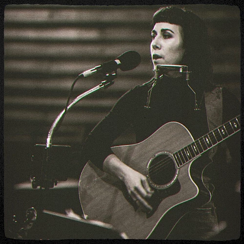 A black and white photo shows a person playing an acoustic guitar and singing into a microphone while also wearing a harmonica holder.