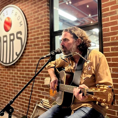 A man is playing a guitar and singing into a microphone in front of a brick building with a sign that says "Ciders From Mars."