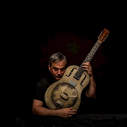A man holding a metal-bodied resonator guitar against a black background, appearing pensive or contemplative.