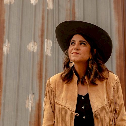 A person wears a brown fringed jacket and a wide-brimmed hat, standing against a rustic metal wall with a thoughtful expression.