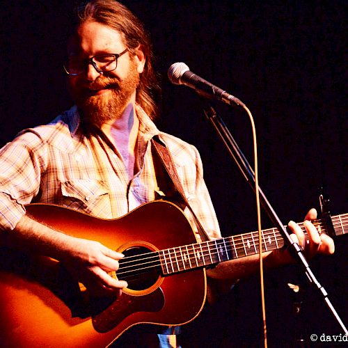 A bearded man is playing an acoustic guitar and singing into a microphone on stage, wearing glasses and a plaid shirt.