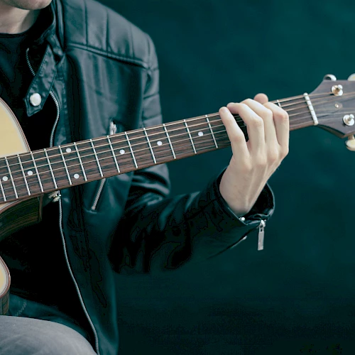A person wearing a black leather jacket is playing an acoustic guitar, focusing on the neck and frets of the guitar.