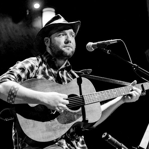 A man wearing a hat and plaid shirt, playing an acoustic guitar and singing into a microphone on stage in a black and white photo.