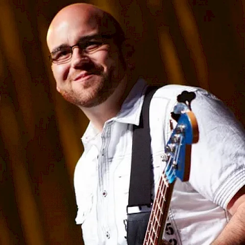 A bald man with glasses, wearing a white shirt, is smiling and holding a blue electric bass guitar against a dark background.