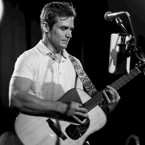 A man is playing an acoustic guitar on stage with a microphone stand in front of him, captured in a black and white photograph.