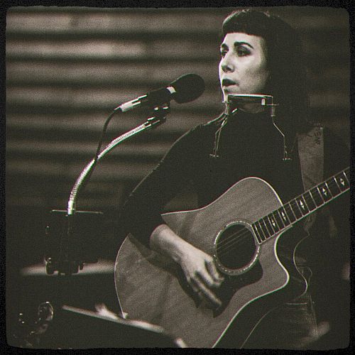 A person is playing an acoustic guitar and singing into a microphone, with a harmonica holder around their neck, in a black and white photo.