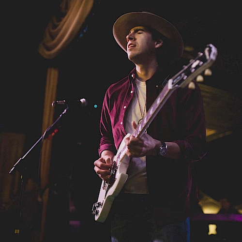 A person in a hat is playing an electric guitar on stage, standing by a microphone. The background is dim with a few lights visible.