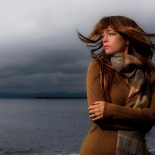 A woman with long hair stands by a body of water, wrapped in a scarf, with wind blowing and dark clouds overhead.