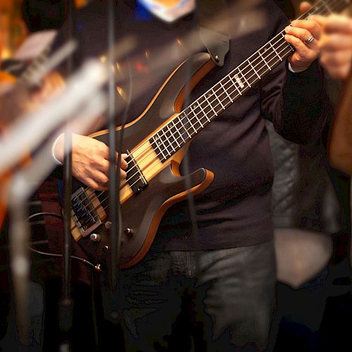 A person playing a bass guitar during a live performance, surrounded by other musicians and equipment in a dimly lit setting.