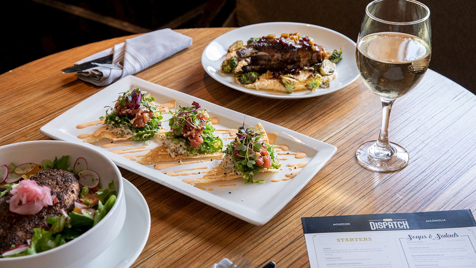 A dining table with a salad, two plated dishes, a glass of white wine, and a menu on a wooden table.