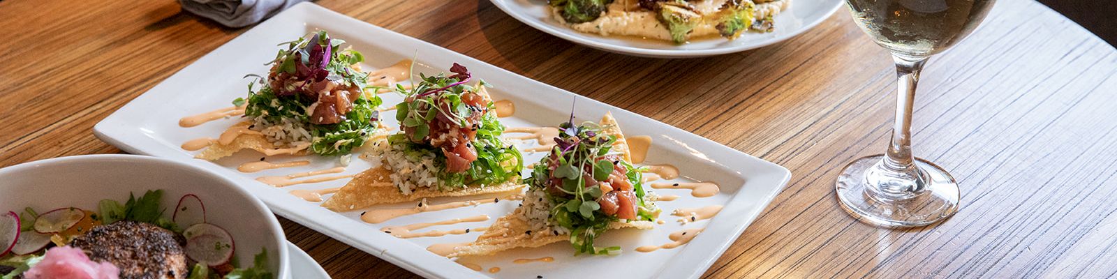 A dining table with a salad, two plated dishes, a glass of white wine, and a menu on a wooden table.