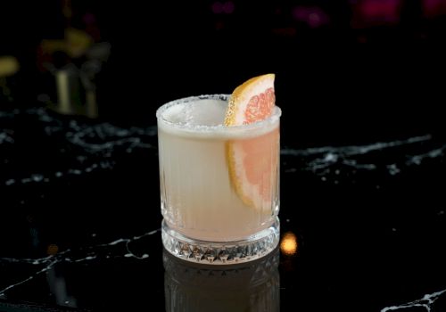 A short, pale cocktail in a thick glass with a salted rim and a grapefruit slice garnish on the rim.