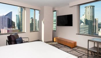A modern room features large windows with a city view, a bed, a wall-mounted TV, a chair, a table, and a wooden bench.