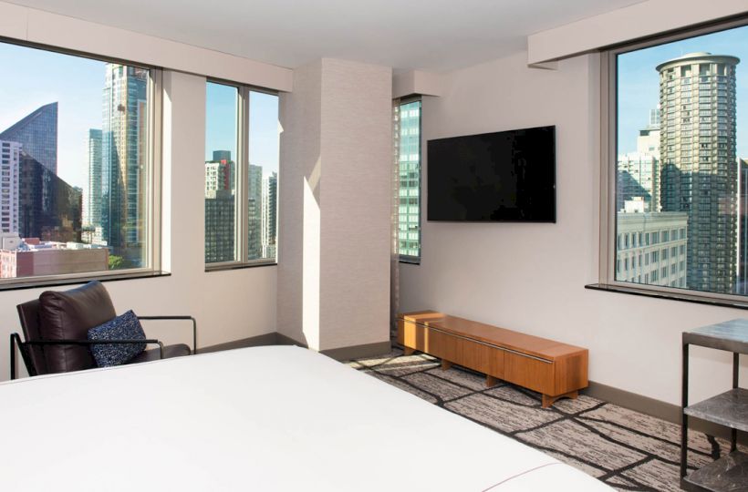 A modern room features large windows with a city view, a bed, a wall-mounted TV, a chair, a table, and a wooden bench.