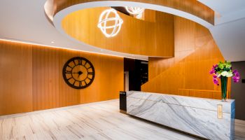 A modern lobby with a marble reception desk, wooden wall decor, a clock, spiral staircase, and hanging light fixtures, plus a vase of flowers.