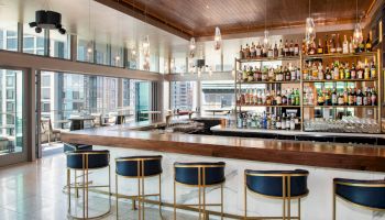 A modern bar features a long counter with blue stools, a wide selection of alcohol behind, pendant lights, and large windows overlooking a cityscape.