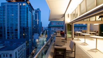 A cityscape view from a balcony with modern buildings, illuminated interiors, and some tables and chairs placed along the open area.