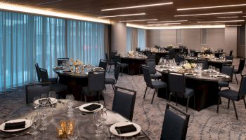 A modern, elegantly decorated banquet hall with round tables set for an event. The tables have black napkins and flowers, with large windows and drapes.