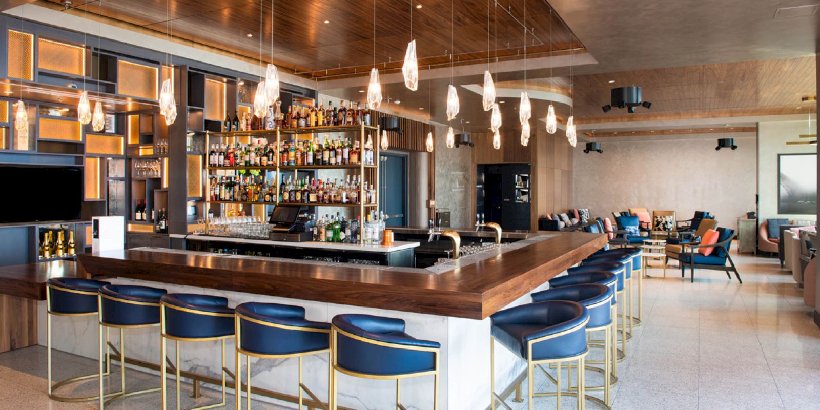 A modern bar with a wooden countertop, blue stools, shelves of liquor, pendant lighting, and seating areas in the background.