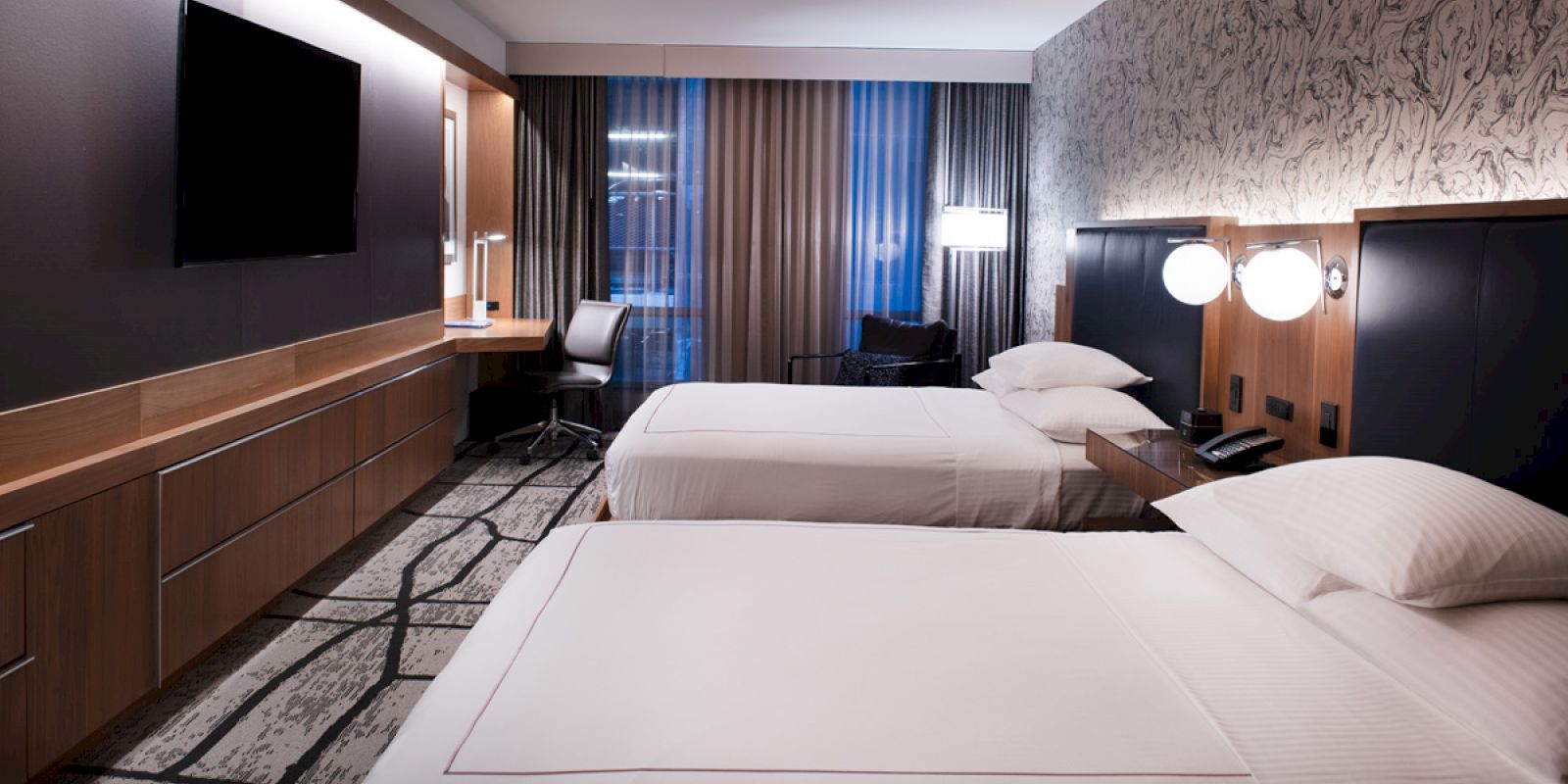This image shows a modern hotel room with two neatly made beds, a large TV on the wall, a desk and chair, and stylish decor accents.