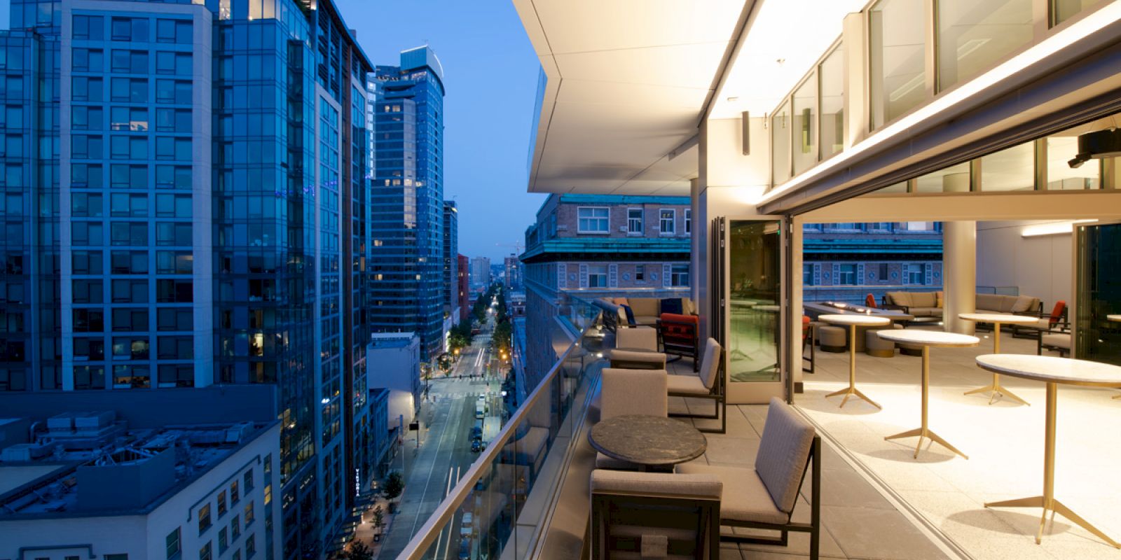 A modern cityscape during dusk with tall buildings, a street view below, and an upscale outdoor patio of a high-rise building featuring tables and chairs.