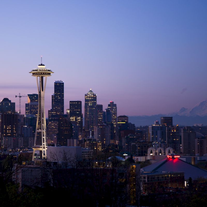 A twilight view of the Seattle skyline featuring the Space Needle with Mount Rainier in the background and city lights starting to twinkle.