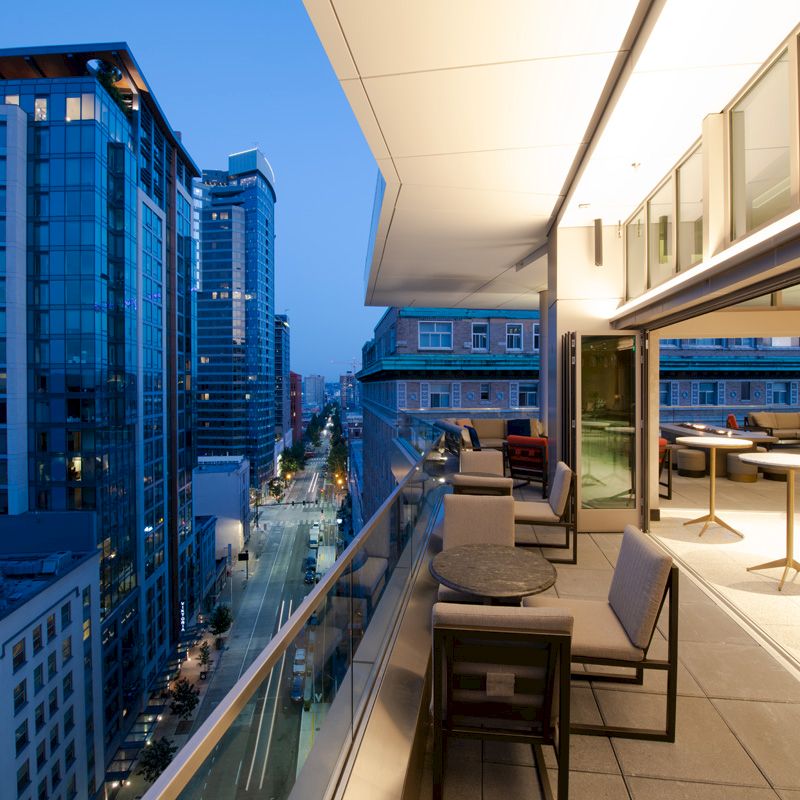 A modern balcony overlooking a cityscape with tall buildings, featuring outdoor seating, tables, and large windows, in the evening.